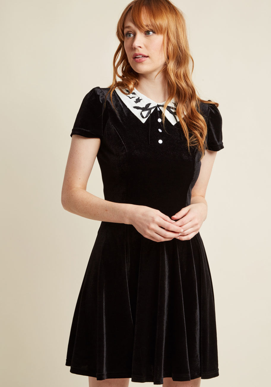 4746 - It takes courage to commit to a look as chic as this black velvet dress by Hell Bunny! The ivory collar - embroidered with a cloud of bats - and pairing of a ribbon tie with pearly decorative buttons on this short-sleeved frock look hauntingly haute along