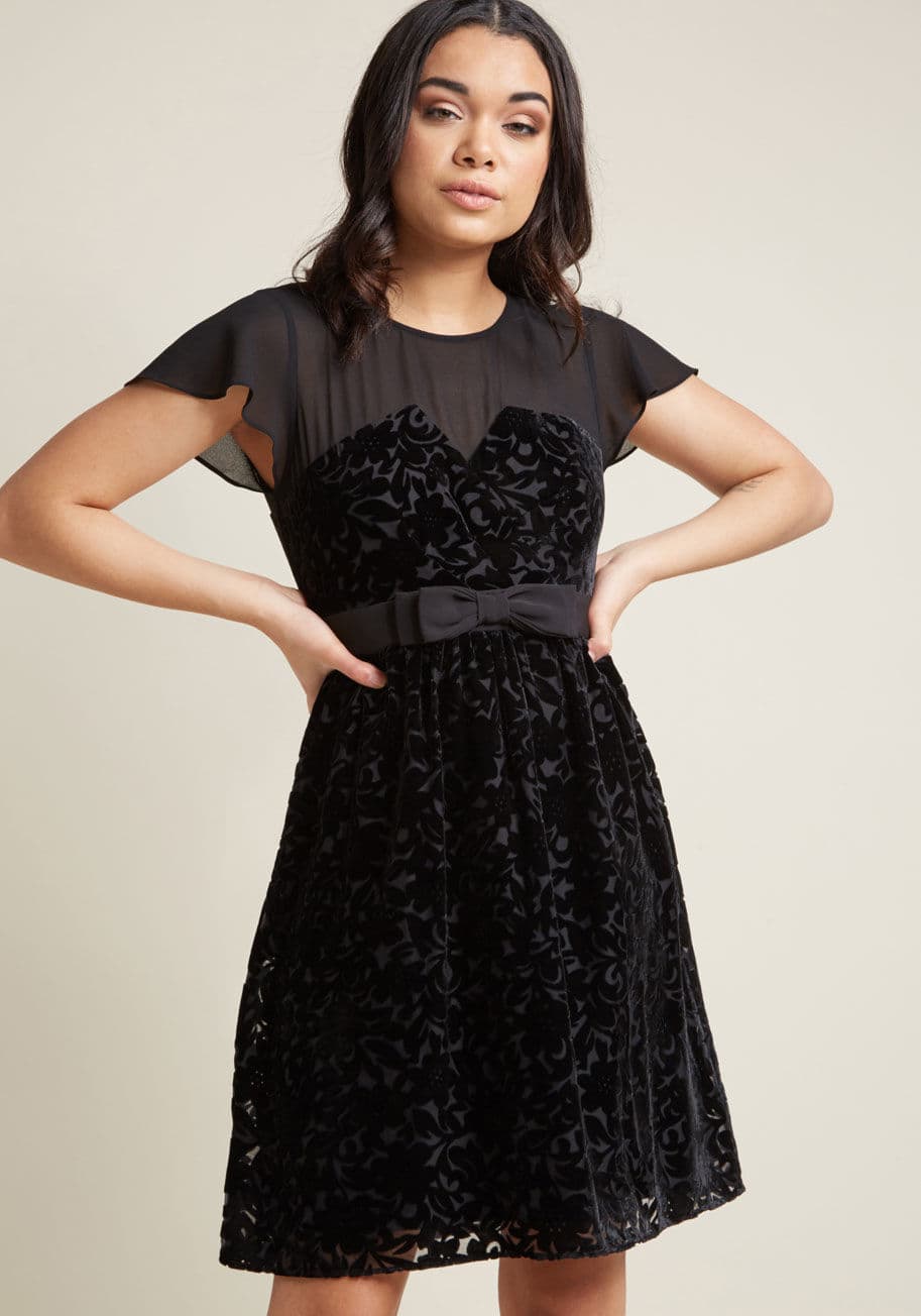 D61410 - This little black dress is a daring and dapper way of achieving the luxury you deserve! A ModCloth exclusive, this fancy A-line features fluttering sleeves and a matching mesh neckline, a notched bust, a bow-topped waistband, and beautiful burnout velvet 