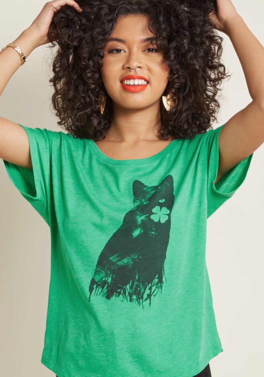 St. Patrick's Cat - This St. Patty's Day, you're ready to welcome all the good luck charms that may come your way! This kelly green T-shirt's black graphic features a painted cat who found himself a festive talisman of his own. With a wide neckline and dolman short sleeves, 